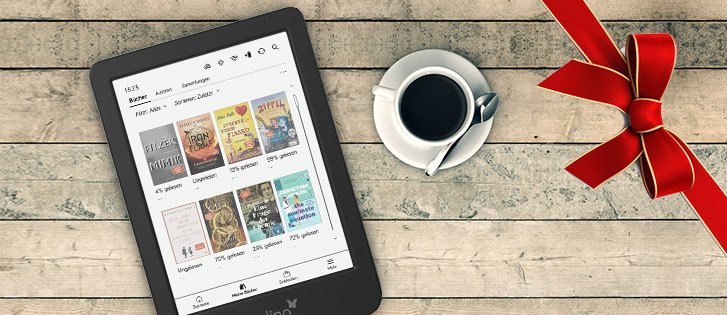 Ein eReader aus der Vogelperspektive gezeigt liegt mit geöffneter Startseite, auf der mehrere Cover zu sehen sind, neben einer gefüllten Kaffeetasse. Rechs im Bild ist eine Geschenkband-Schleife zu sehen.